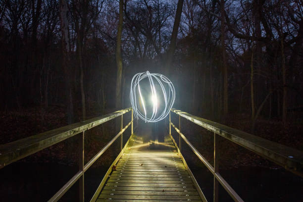 Enveloped by darkness, a wooden footbridge in a forest comes alive at night. A mesmerizing light painting creates a guiding orb, illuminating the path through the shadows. This captivating image captures the mystery and creativity of the night.