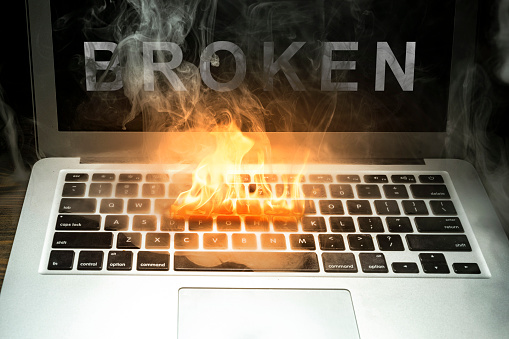Close up laptop burned with flame and smokes on a wooden desk with broken word isolated over black background. Broken equipment.