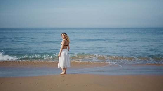Woman relaxing ocean shore walking on calm foamy waves alone. Relaxed curly model strolling on wet sand looking beautiful seascape. Carefree romantic female tourist enjoy sunny summer weekend.