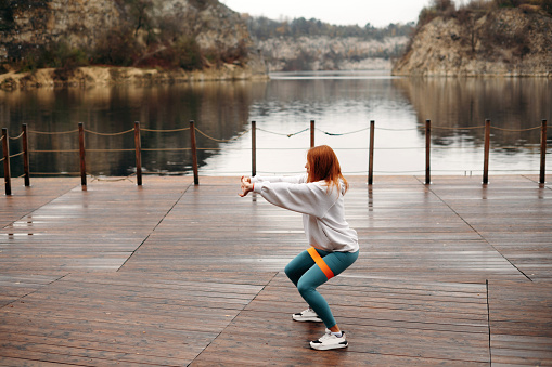 Stylish sporty fit woman with red hair in sportswear squats with fitness elastic band on wooden pier near the lake with rocks. Sports concept, healthy lifestyle