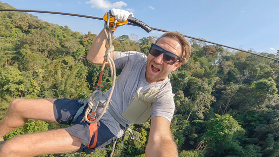 Amidst the vibrant Laotian jungle, a young man finds exhilaration zip-lining through the lush canopy, captivated by the stunning natural scenery below.
