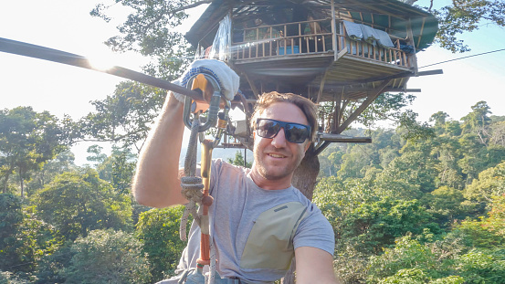 Amidst the vibrant Laotian jungle, a young man finds exhilaration zip-lining through the lush canopy, captivated by the stunning natural scenery below.