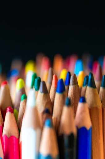 Color image depicting a close up macro view of a collection of colored pencils, on a black background. Selective focus image with one of the pencils in sharp focus with the others blurred out of focus.