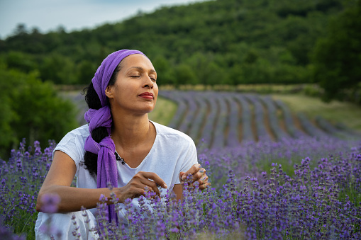 A Latina woman with a purple headscarf and a hat is holding a bunch of lavender in the middle of a lavender field at sunset.