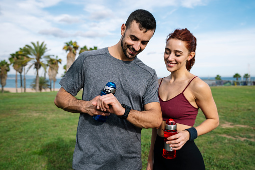 Runners resting after training looking at sports watch checking heart rate. Joyful fit couple jogging in park checking smartwatch while holding water bottle outside.