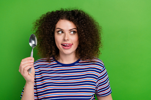 Portrait of creative cute person tongue lick teeth hand hold spoon look empty space isolated on green color background.