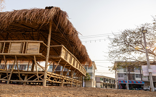 Close-up low angle view of bamboo shed huts with thatched roofs installed on a mound near residential buildings and a paved road in rural Thailand.