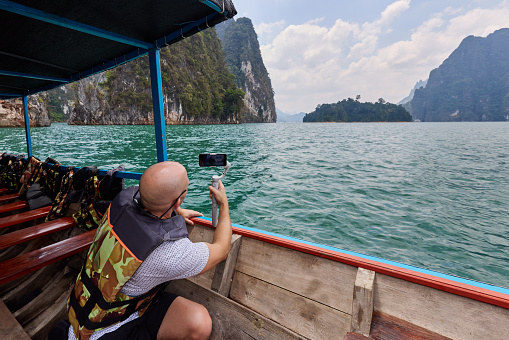 A man uses his smartphone on a boat to film the islands in the reservoir in Khao Sok National Park in Thailand