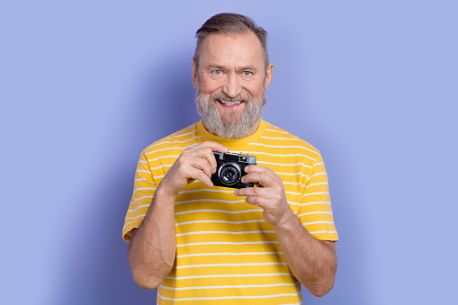 Photo of toothy beaming senior man with gray beard dressed striped t-shirt holding vintage camera isolated on purple color background.