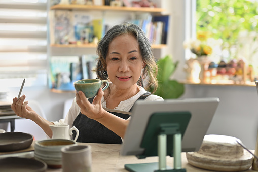 Senior Artist or start up small business owner selling a handcraft of ceramic coffee cup on an online live steam via social media platform on tablet.