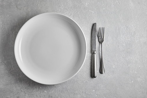 Empty white ceramic plate and fork and knife on gray stone table. Top view with a copy of the space.