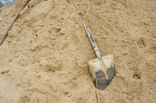 A worn-out shovel lies in a pile of sand at a construction site, suggesting ongoing work. An abandoned shovel on a large sand heap, indicating a pause in labor.