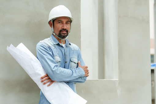 Multiethnic construction engineers wearing hard hats stand with folded arms holding structural diagrams. Standing on ivory leaning against the wall of building Supervise construction work, build house