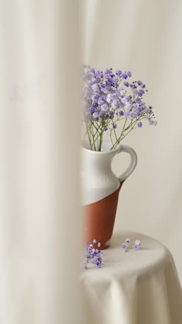 White curtain partially open waving next to a jar with purple flowers