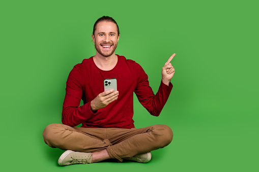 Full length photo of cheerful man dressed pullover sit hold smartphone indicating at offer empty space isolated on green color background.