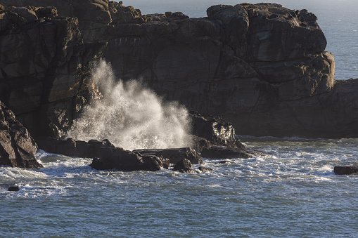 A rocky shoreline with a large wave crashing into the rocks. The water is calm and the sky is clear