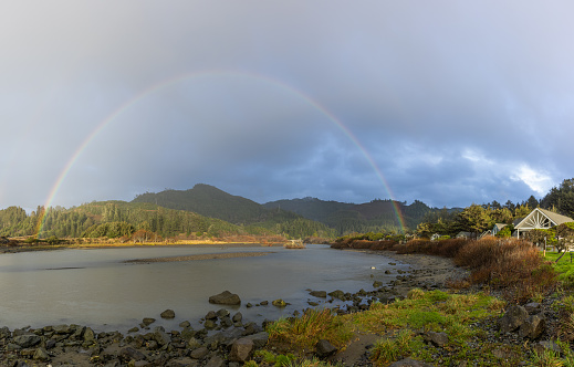 A rainbow is seen over a body of water with a house in the background. The sky is cloudy and the water is calm