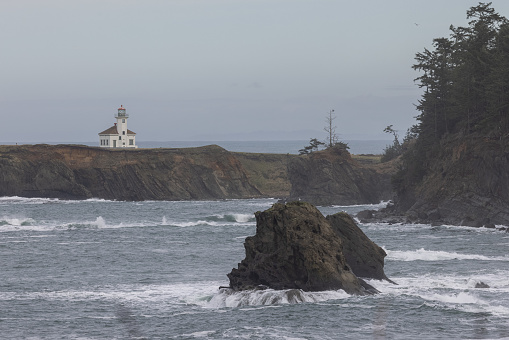 A lighthouse is on a rocky shoreline. The water is choppy and the sky is overcast