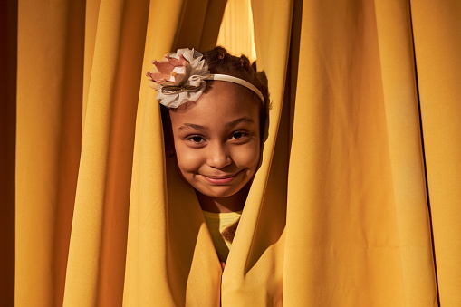 Close up portrait of smiling Black girl peeking through yellow curtains on stage copy space