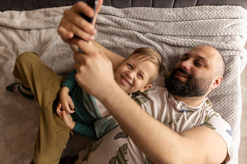 Dad and little son laughing looking at cell phone photo. They are lying on the bed in the bedroom and taking photos. A father hugs his son. They make funny faces. A man with dark hair and a strong beard and a boy with light hair and eyes