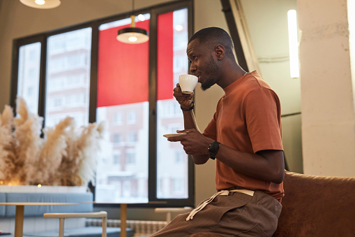 Side view portrait of Black adult man enjoying cup of coffee in cafe or office setting with low light copy space