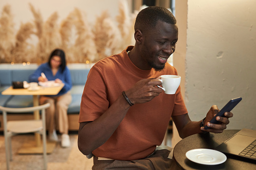 Side view portrait of smiling African American man using smartphone in coffee shop and enjoying cup of coffee cozy low light copy space