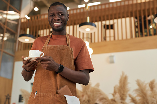 Waist up portrait of smiling Black man as barista holding coffee cup in cafe and wearing apron, copy space