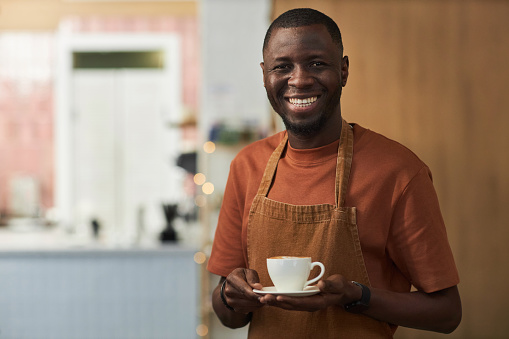 Waist up portrait of smiling African American man as cafe worker holding coffee cup and wearing apron, copy space