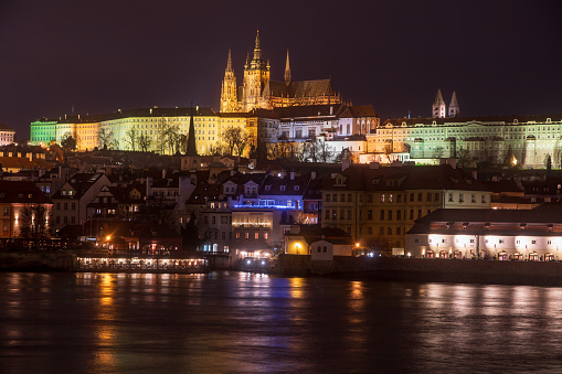 Image of St Vitus Cathedral, Prague taken at night from river level. The buildings are illuminated and the lights are reflected on the surface of the water. The shot is a long exposure which has smoothed out the surface of the water and created starbursts of the lights.