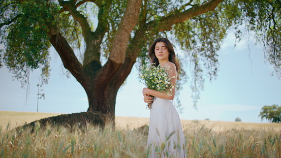 Young woman holding flowers at rye field. Tender lady enjoying rural nature carrying bouquet of chamomiles. Beautiful serene girl in long white dress looking camera posing at huge tree background