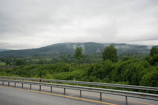 Norwegian mountains, fjords with green trees surrounded by white fog on a cloudy, rainy day with a highway in the foreground.