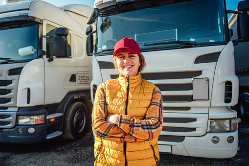 Portrait of a smiling female truck driver standing in front of a truck vehicle in the parking lot. Transportation service