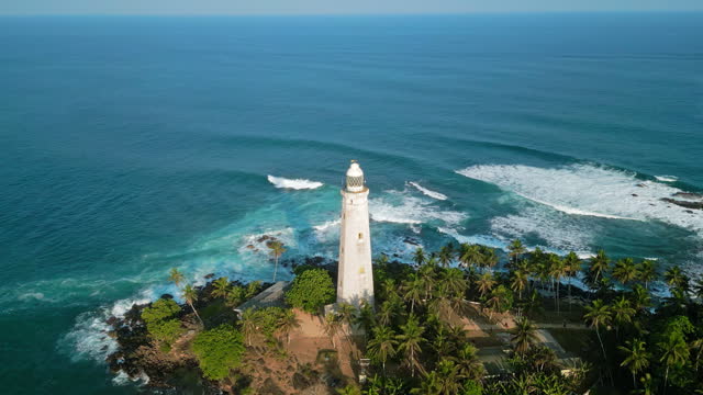 Aerial view vintage Dutch lighthouse on tropical island, surrounded by greenery, azure ocean waters. Waves crash against rocky coast guiding vessels through bay. Drone navigation, safety.