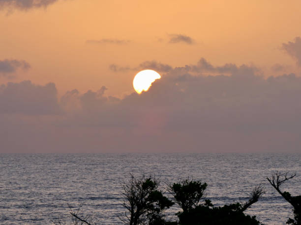 A bright yellow sun emerges from behind pinky lavender clouds on a golden backdrop on the horizon of the Atlantic Ocean along the coast of St. Maarten.
