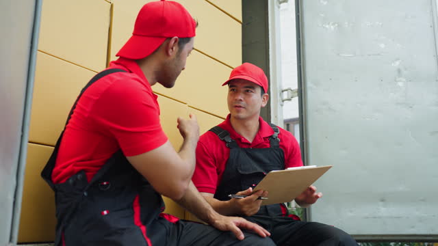 Delivery man sit and discuss together using document in clipboarad and stay on back part of truck during move package or boxes to customer house.