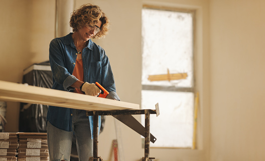 Woman wearing safety goggles is indoors, using a crosscut saw to trim a wooden plank for a home renovation project. Her skilled woodworking and dedication to upgrading the house are evident in this DIY endeavor.