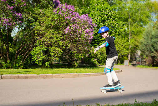 A 10-year-old boy rides a plastic skateboard in the park on a sunny day. a child in a protective helmet, with protection on his arms and legs, rides a skateboard against the backdrop of blooming lilacs on a sunny spring day.