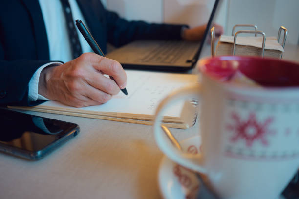 istock Close-up of a man in suit sitting at table with laptop writing on a notebook 2081617214