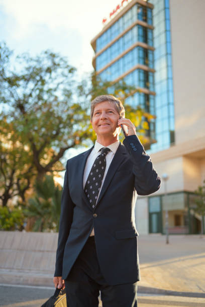 A man dressed in a formal suit is engaged in a phone conversation while standing in an urban setting. He appears focused and professional with a business building in the background