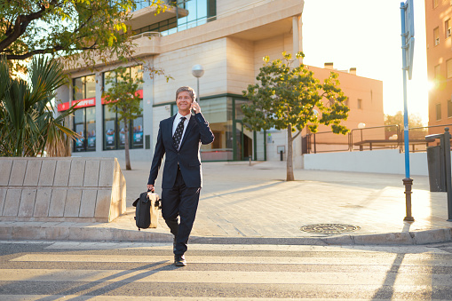 A man dressed in a formal suit is engaged in a phone conversation while standing in an urban setting. He appears focused and professional with a business building in the background