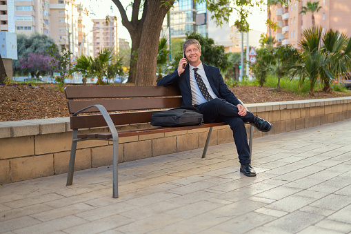 A man is seated on a bench, engaged in a phone conversation. He is holding a cell phone to his ear while talking. The background shows a park setting with trees and business buildings.