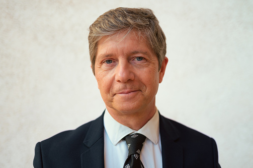 A professional man dressed in a suit and tie is standing and posing for a portrait, appearing confident and well-groomed. The background is neutral, drawing attention to his attire and demeanor.