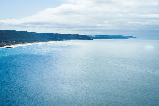 The blue waters of the Atlantic Ocean and the coastal sandy strip and mountains, view from above. A place near Nazare, Portugal. Image in blue tones and blue haze in the distance.