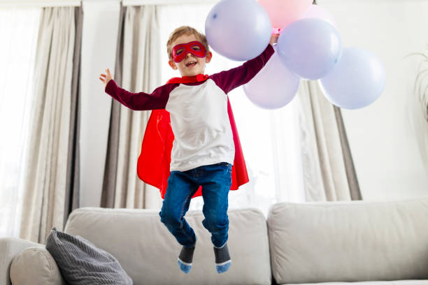 istock Boy in superhero costume with balloons jumping on sofa 2081629395
