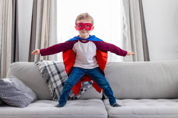 istock Boy in superhero costume with cape and mask standing on sofa 2081629922