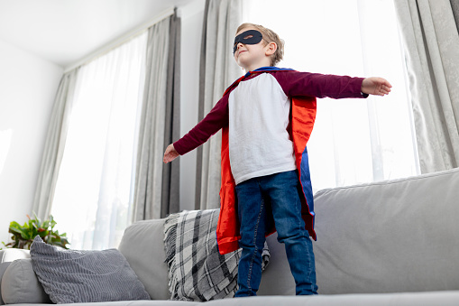 Young child imagines being a superhero, standing confidently in a living room with a cape.