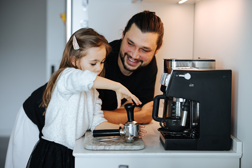Father and daughter at home using technology home virtual assistant device in family kitchen. Girl smelling coffee prepared by father at home