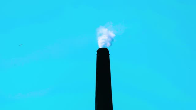 Static shot of a tall factory chimney releasing smoke at blue sky background. Closeup.