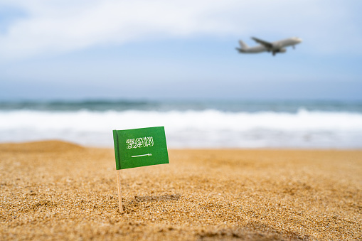 Flag of Saudi Arabia in the form of a toothpick in the sand of beach opposite sea wave with landing airplane. Travel concept