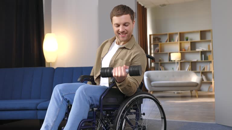 Physical rehabilitation for disabled people. Happy guy in wheelchair making exercises with dumbbell at home. Cheerful impaired young man working out with weights indoors